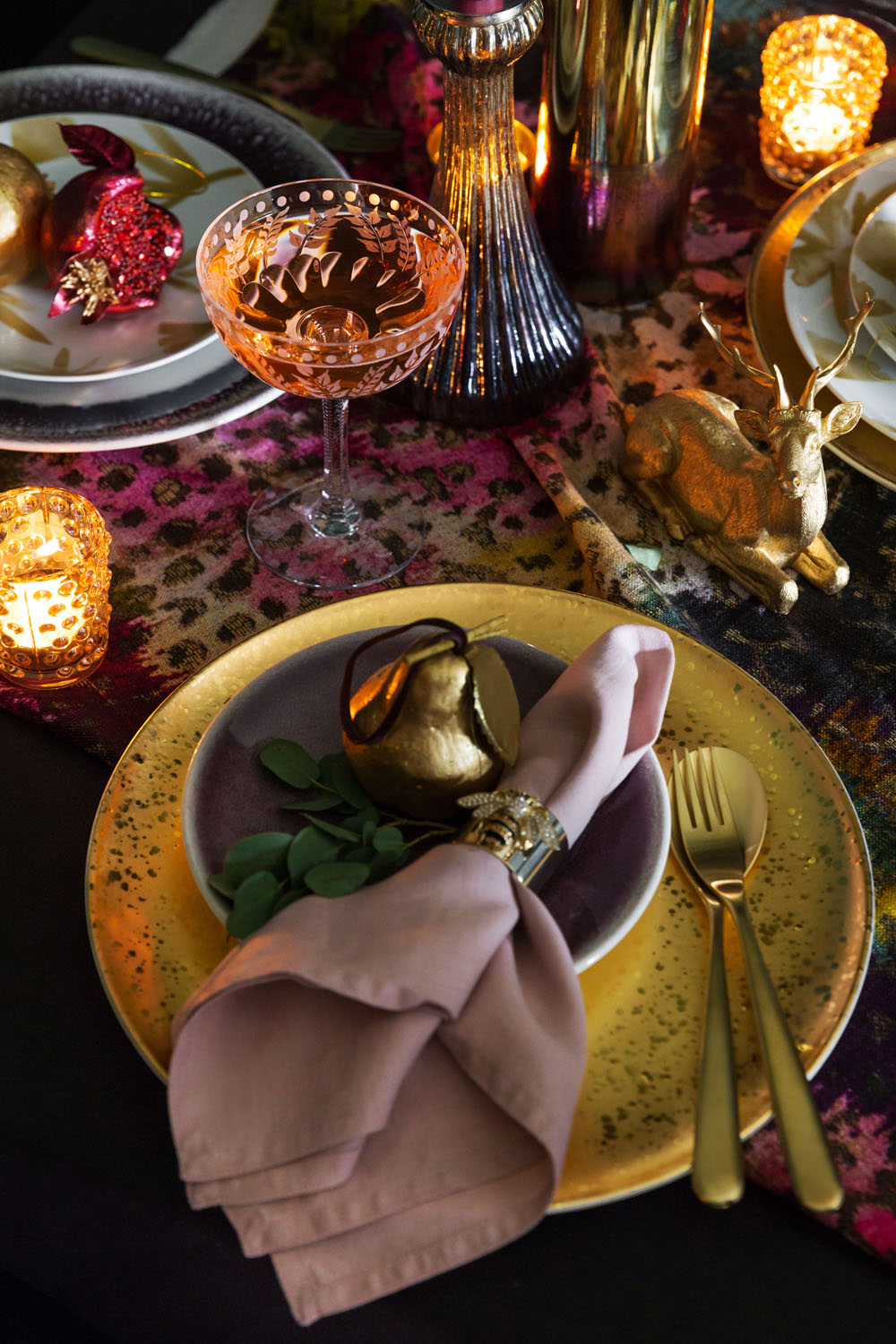 A festive table setting with a gold-rimmed plate, a mauve serviette with a gold serviette ring, gold cutlery, a golden pear ornament, a sprig of greenery, and lit candles on a patterned tablecloth.