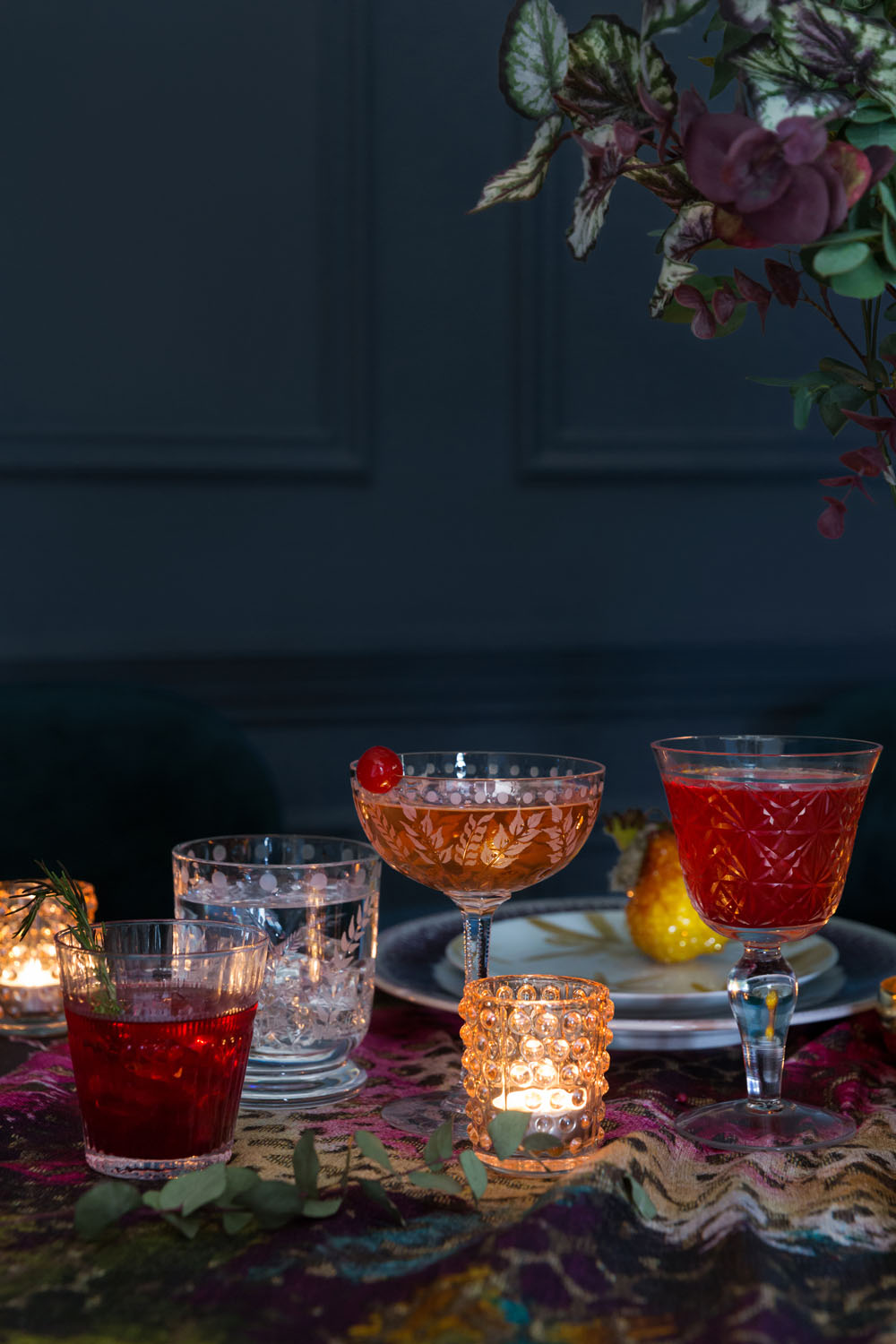 A table with three cocktails in vintage glasses, a small glass candle holder, another candle in the background, and a dark floral tablecloth. Dim lighting and a floral arrangement are partially visible at the top right.