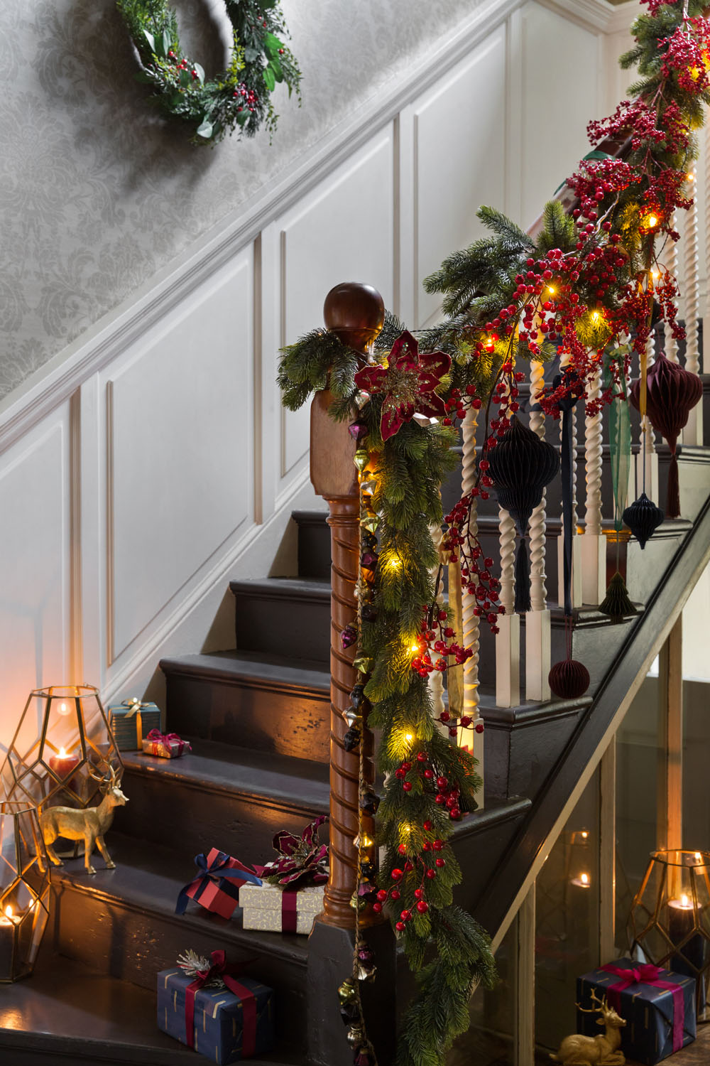 A staircase decorated with garlands of greenery, red berries, baubles, and fairy lights. Wrapped presents and gold holiday decorations are on the steps and floor. A wreath hangs on the wall above the staircase.