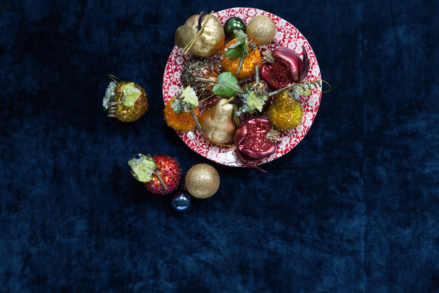 A red and white patterned plate holds assorted holiday baubles, including gold, red, and green pieces shaped like fruit and hearts, on a dark blue textured surface. Some baubles lie scattered beside the plate.