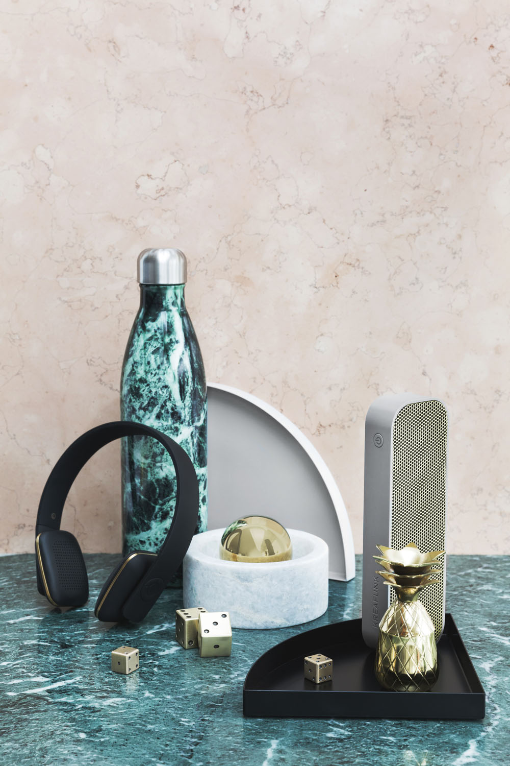 A marble water bottle, wireless headphones, portable speaker, gold pineapple container, gold ball, dice, and modern trays are displayed on a green marble surface with a light marble background.