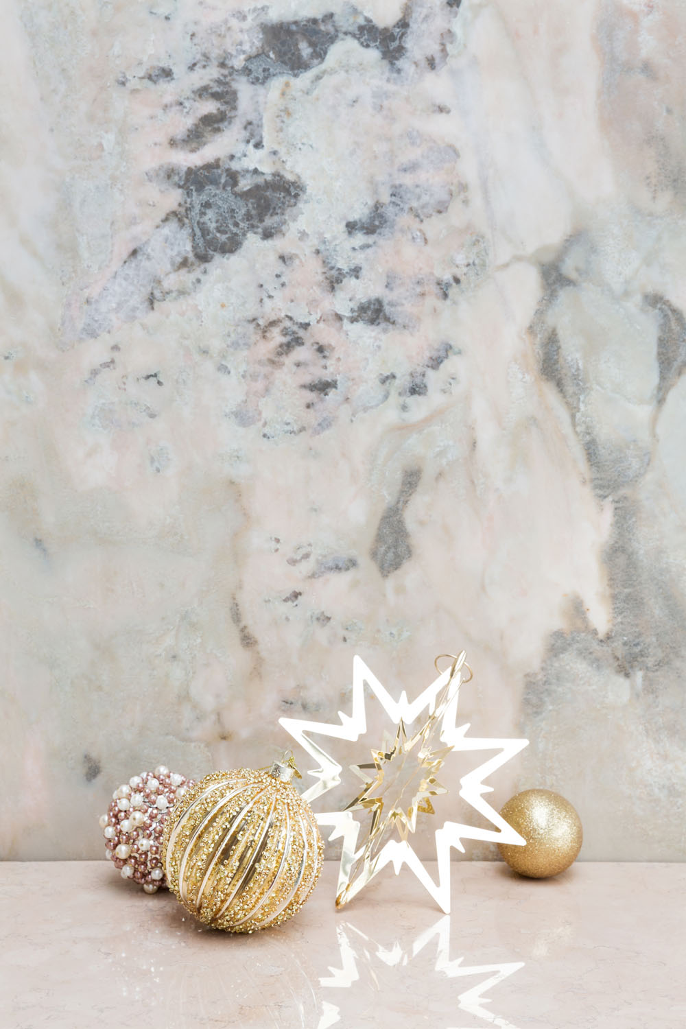 Three Christmas baubles and a star decoration are arranged on a light marble surface with a marble wall background. The baubles include a gold ball, a gold striped ball, and a beaded ball.