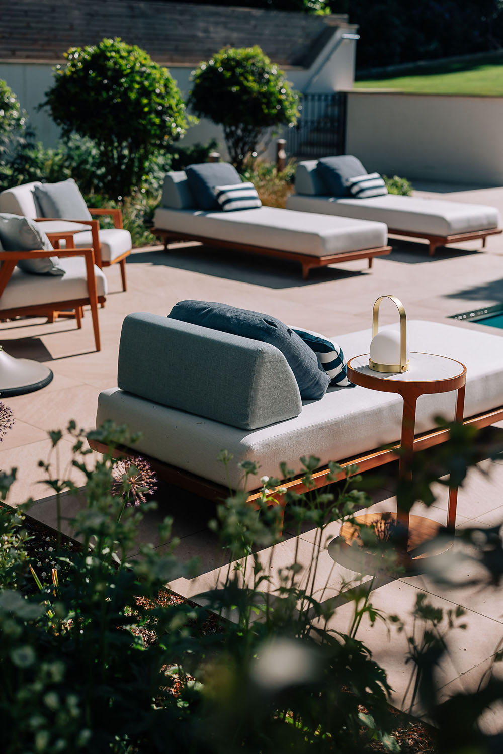 Outdoor patio with cushioned lounge chairs, striped pillows, and small wooden side tables. A portable lamp is on one table. Sunlight and shadows fall across the area, with plants and greenery in the foreground and background.